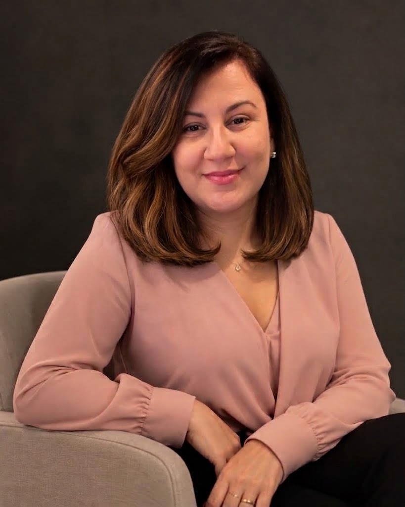 Luciana Borges, psicanalista, sentada em uma poltrona clara, vestindo blusa rosa, com fundo escuro